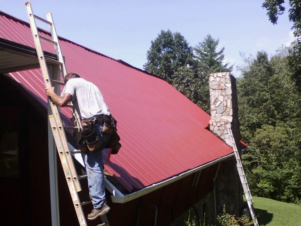 Roofer on a ladder beside a red metal roof and stone chimney — Soffit & Fascia Repair by Roofing Brothers in Plainville